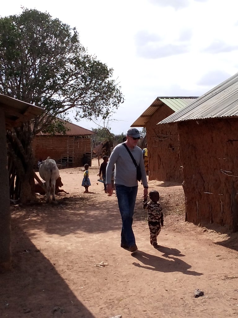 A man walking hand in hand with a young child through a rural Kenyan village, surrounded by simple homes and farm animals, symbolizing community connection and care.